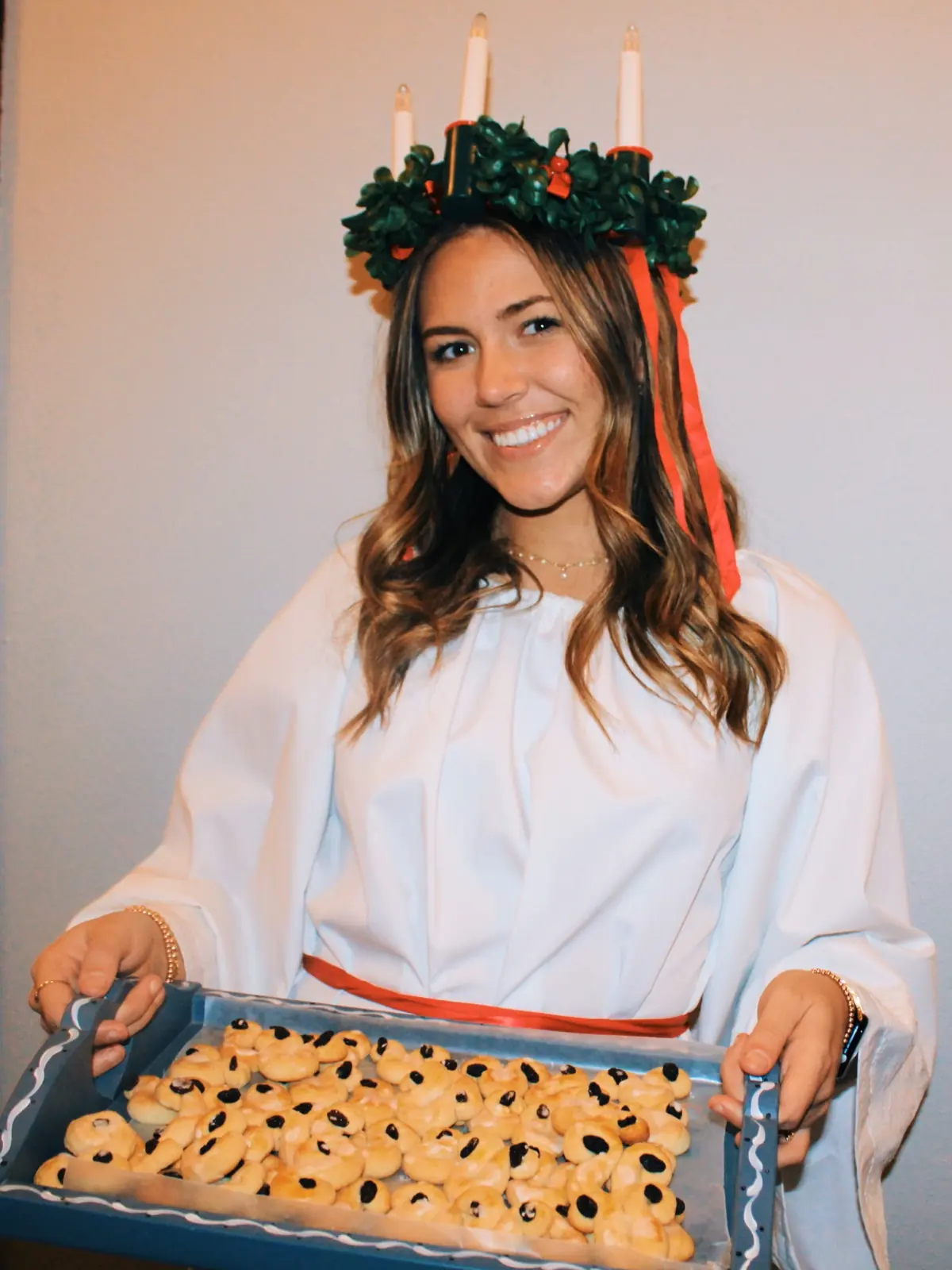 Young woman in a white Lucia gown and candle crown holding a tray of fresh-baked treats.
