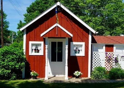 A small red stuga with white trim, lace-curtained windows and flower boxes on the museum grounds.
